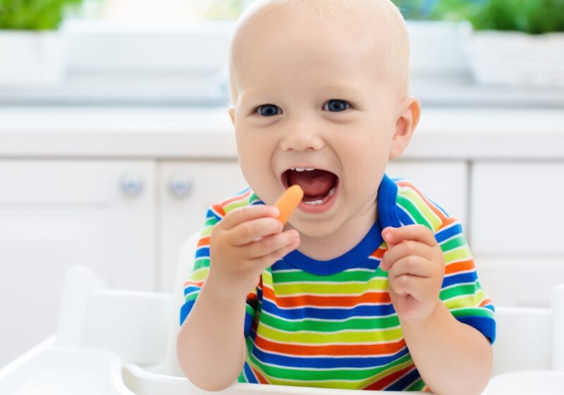 Bebé sorridente a comer cenoura e brócolos frescos numa cadeira de refeição, promovendo uma alimentação saudável e natural